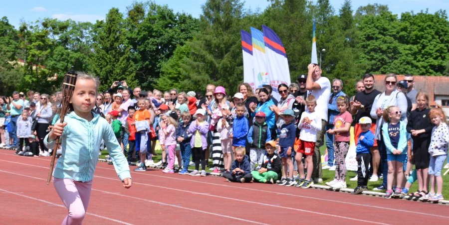 Duża grupa zawodników stoi na stadionie. Po lewej stronie biegnie dziewczynka ze zniczem.