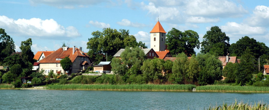 Widok od strony jeziora na wieś. W tle dachy domów i wieża kościoła wśród zieleni.
