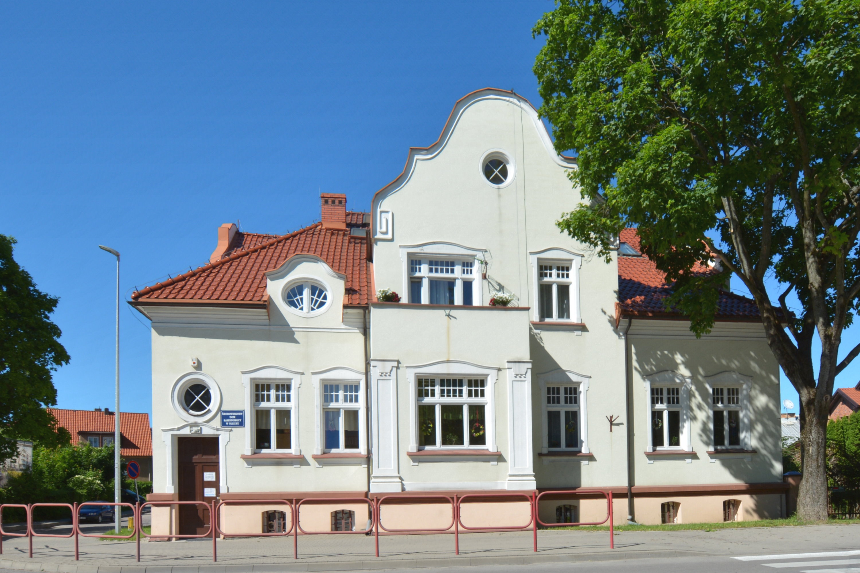 Budynek o zwartej dwukondygnacyjnej bryle przykrytej dachem z czerwonej  dachówki. Po prawej stronie drzewo.