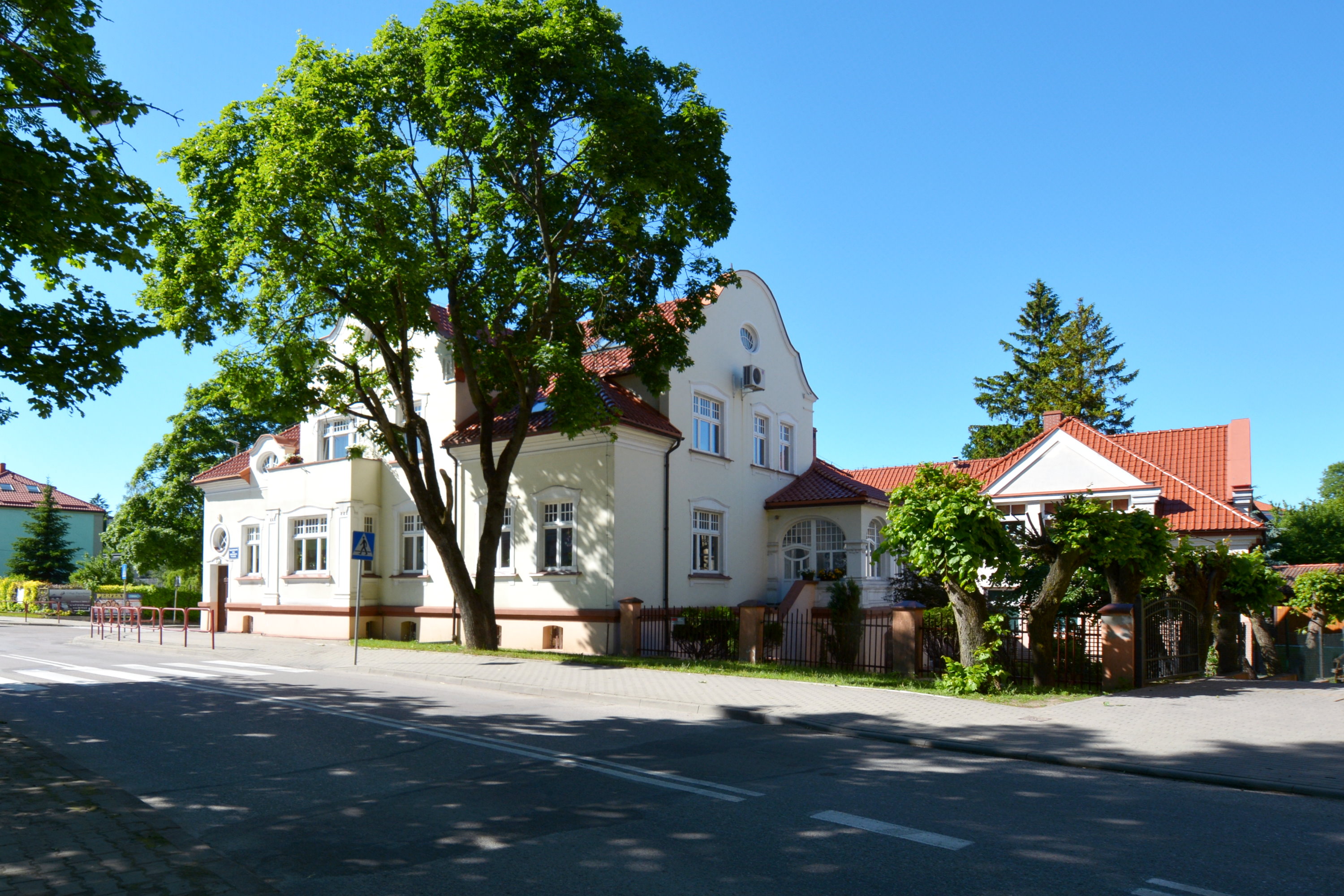 Budynek o zwartej dwukondygnacyjnej bryle przykrytej dachem z czerwonej  dachówki. Po prawej stronie drzewo.