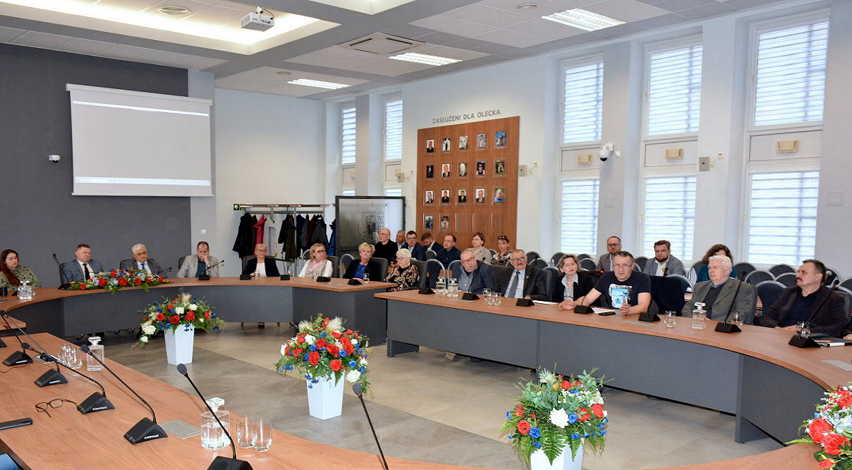 Duża sala z szerokim stołem. Wokół siedzą kobiety i mężczyźni. Dalej okna.