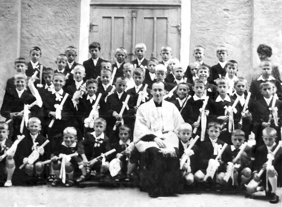 Duża grupa dzieci pozuje do fotografii. W środku siedzi  ksiądz.