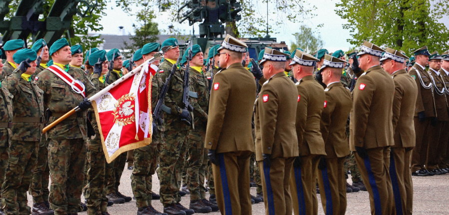 Uroczystość wojskowa – żołnierze w mundurach składają salut. Widoczna jest polska flaga wojskowa oraz żołnierze w zielonych beretach i brązowych mundurach.