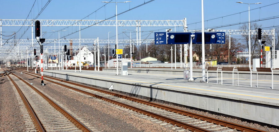 Stacja kolejowa z pustymi peronami, kilkoma torami i sygnalizacją świetlną. Na niebie brak chmur, a w tle widać zabudowania i słupy trakcyjne.