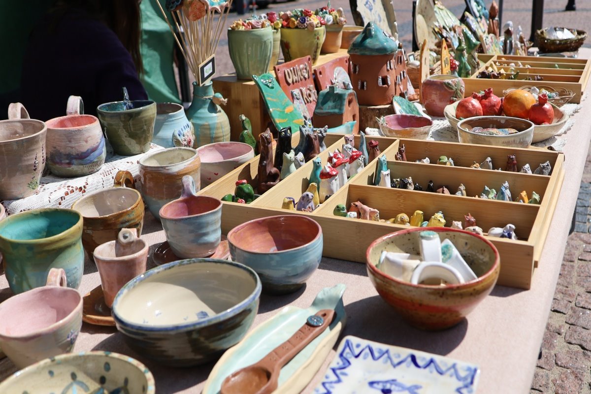 Stoisko z ręcznie robioną ceramiką na targu rzemiosła. Kolorowe miski, kubki, talerze i małe figurki ułożone na stole. Widoczny talerz z motywem ryb i drewniane pudełka z drobnymi wyrobami.
