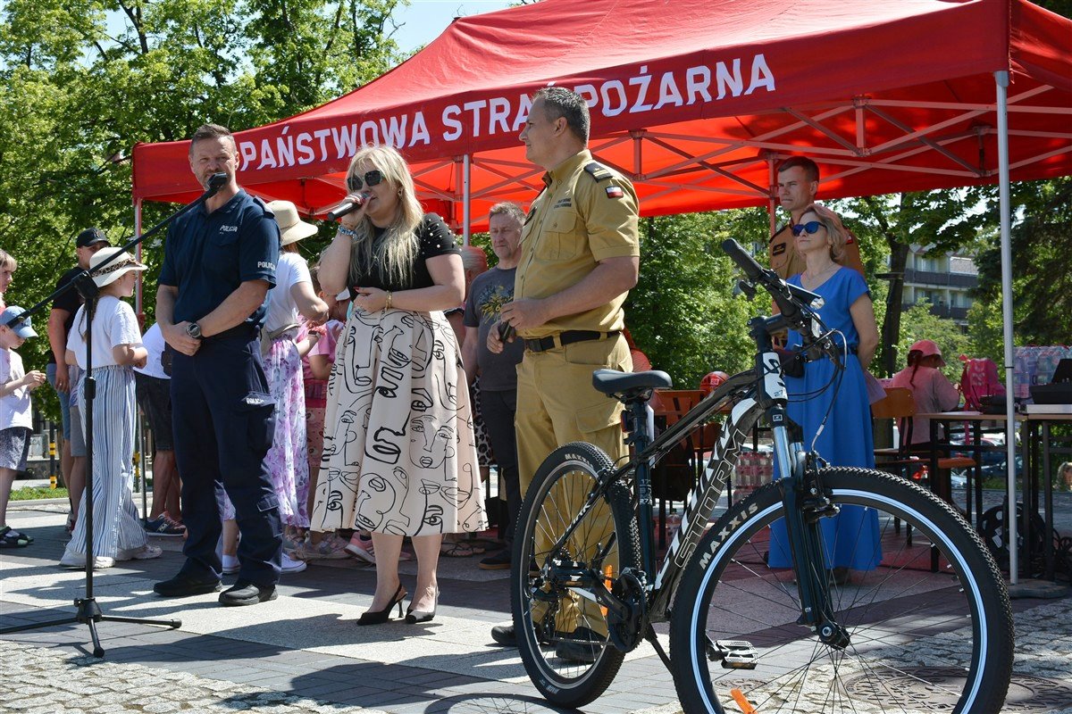 Wydarzenie pod czerwonym namiotem Państwowej Straży Pożarnej. Kobieta przemawia przez mikrofon, obok stoją policjant i strażak. Na pierwszym planie rower górski. W tle widoczni widzowie.