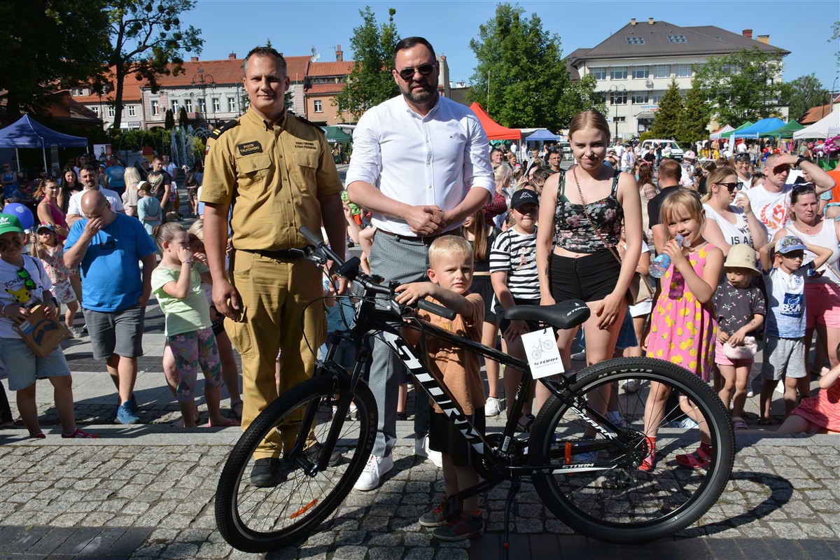 Chłopiec z nowym rowerem górskim, obok strażak w mundurze i mężczyzna w białej koszuli. W tle tłum ludzi na festynie miejskim i kolorowe namioty handlowe.