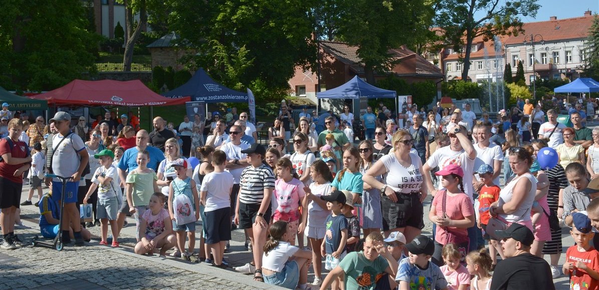 Tłum ludzi, głównie rodziny z dziećmi, zgromadzony na miejskim festynie. W tle kolorowe namioty handlowe i zabytkowe budynki. Słoneczny letni dzień.