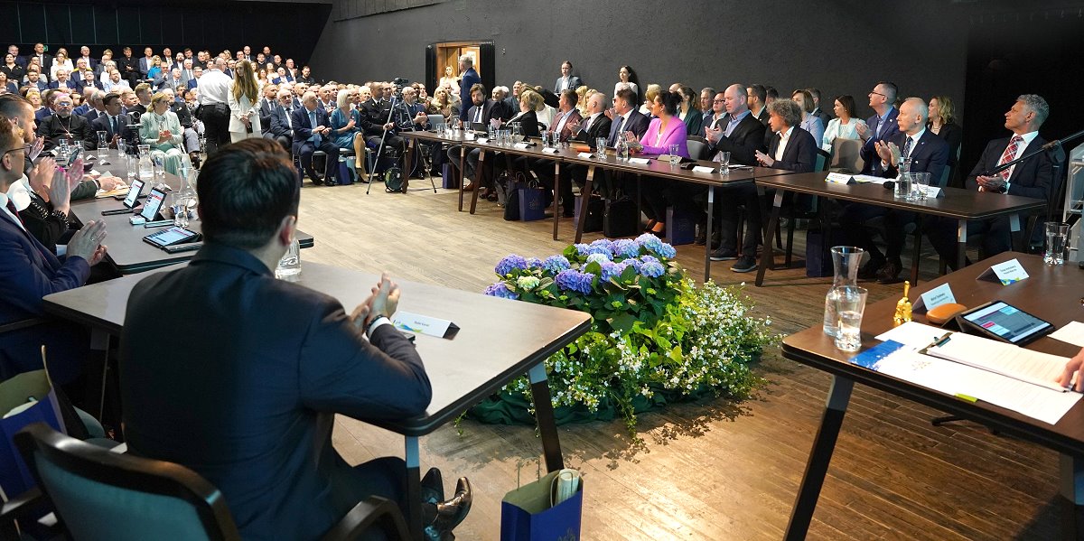 Uczestnicy uroczystej sesji Rady Miasta Ełku siedzą przy stołach ustawionych w podkowę. W tle widownia zapełniona gośćmi. Na środku sali ozdobna kompozycja kwiatowa.