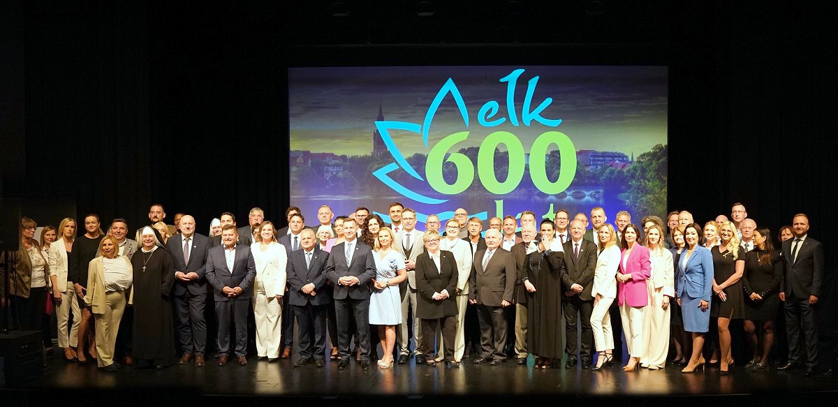 Grupa ludzi pozuje do wspólnego zdjęcia na scenie. W tle widać duży napis „Ełk 600 lat” na kolorowym ekranie. Wszyscy są elegancko ubrani, uśmiechają się i patrzą w stronę aparatu.