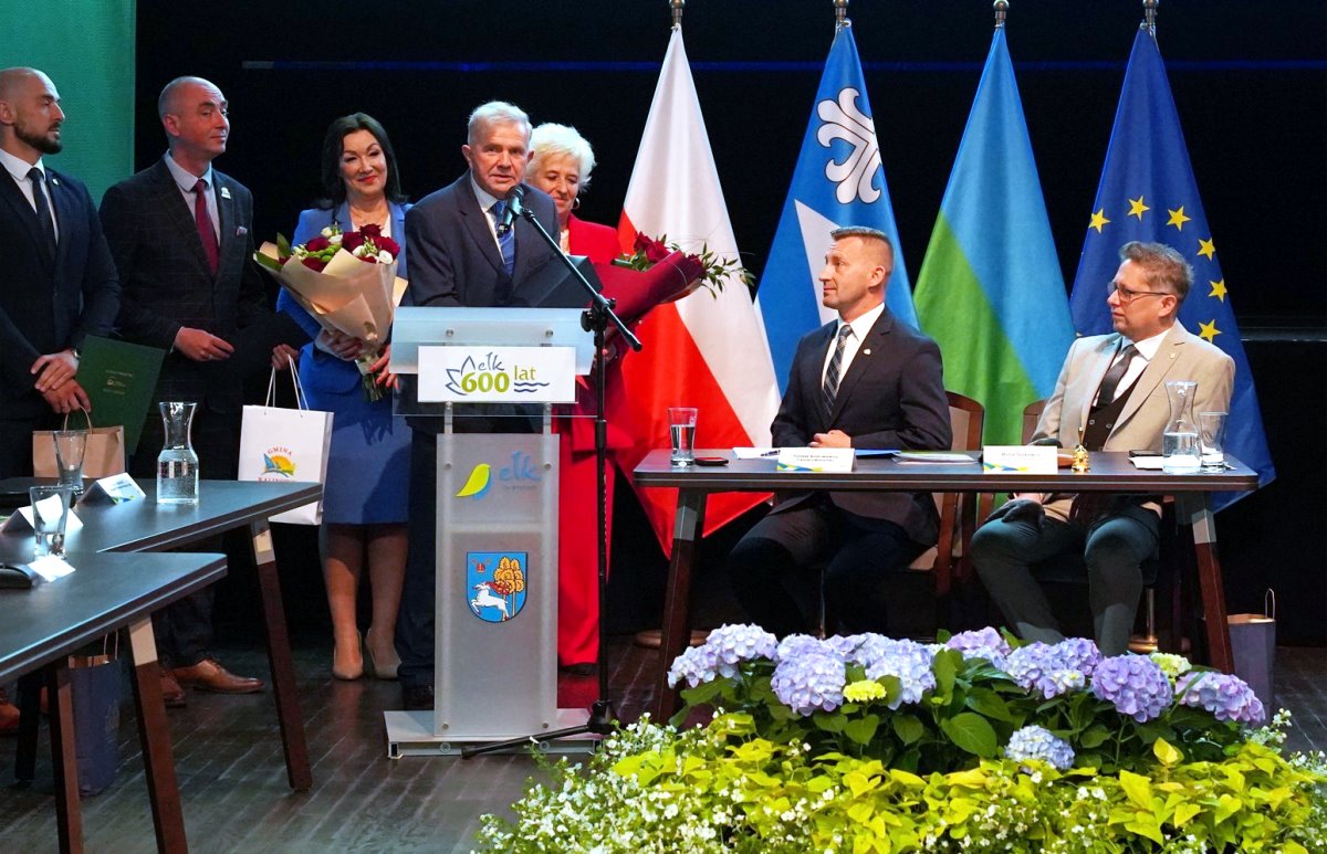 Grupa osób podczas uroczystości z okazji 600-lecia Ełku. Mężczyzna przemawia przy mównicy, obok niego stoją osoby z kwiatami. W tle flagi Polski, Ełku i Unii Europejskiej oraz dekoracje z kwiatów.
