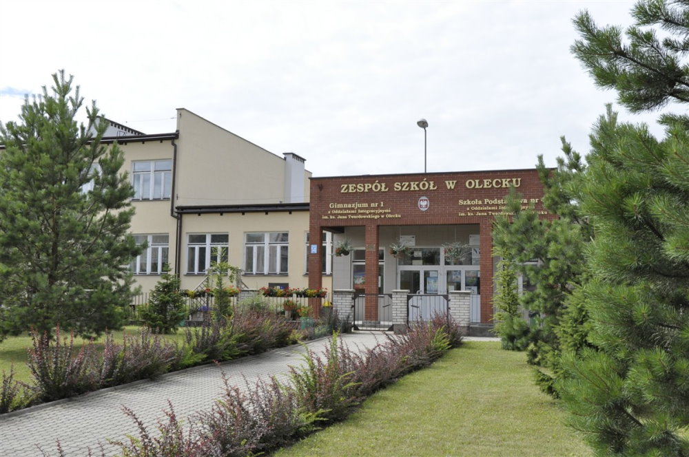 Nowoczesny budynek szkoły w Siejniku z wejściem otoczonym roślinami i drzewkami.