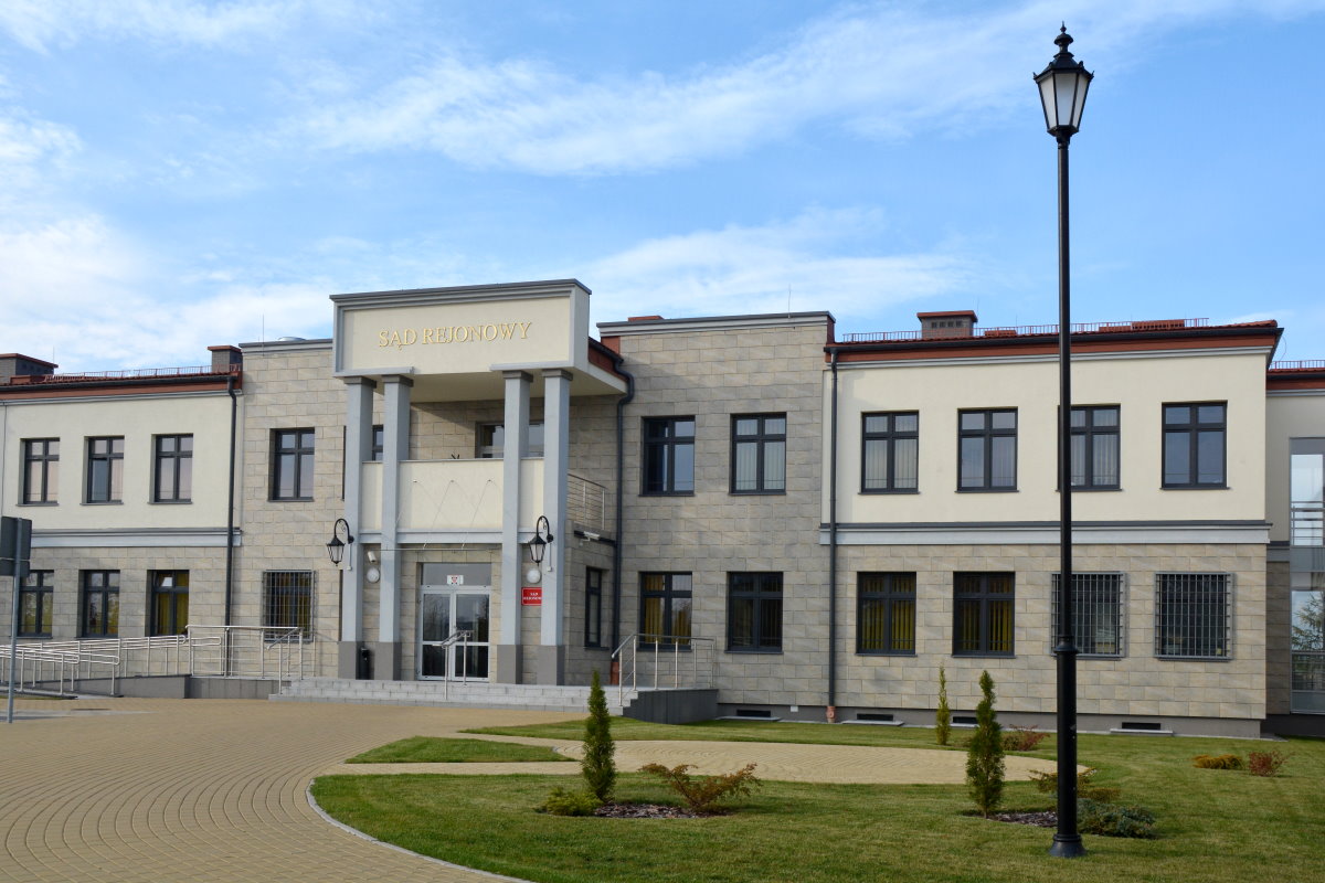 Biały budynek z latarnią przed nim, stojący na placu z chodnikiem i trawnikiem.