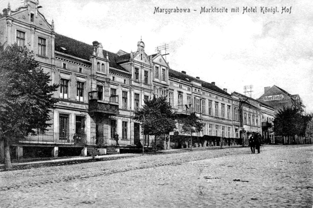 Czarno-białe zdjęcie rynku w Marggrabowej z eleganckimi kamienicami. Widać Hotel Königl. Hof, drzewa przy chodniku i kilka osób spacerujących po brukowanej drodze.