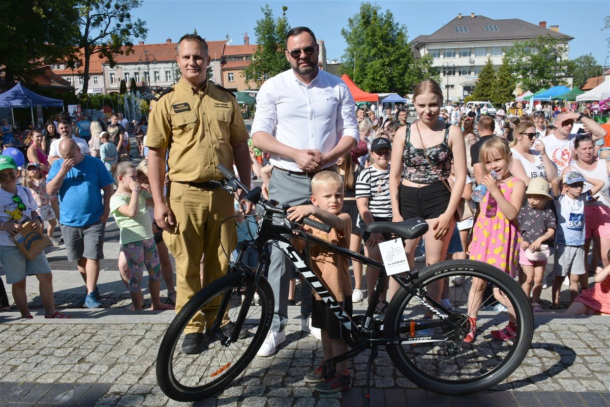Grupa ludzi na miejskim festynie. Na pierwszym planie chłopiec z nowym rowerem, obok niego mężczyzna w mundurze, drugi mężczyzna w białej koszuli i kobieta w letnich ubraniach. W tle tłum i kolorowe namioty.