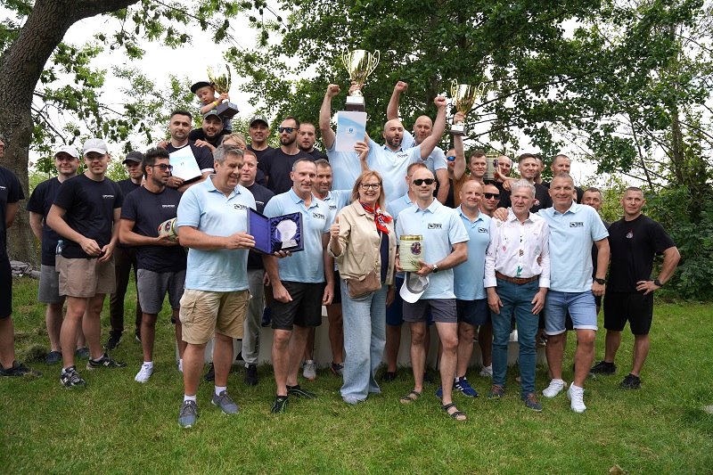 Grupa osób pozuje do zdjęcia na trawie, część z nich trzyma puchary i dyplomy. W tle drzewa. Wszyscy wyglądają na zadowolonych, prawdopodobnie po zakończeniu zawodów lub uroczystości.