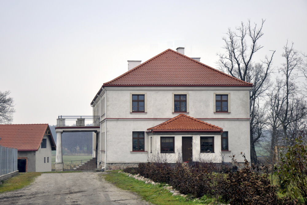 Duży, biały dwór z czerwonym dachem, stoi przy żwirowej drodze, otoczony drzewami bez liści.