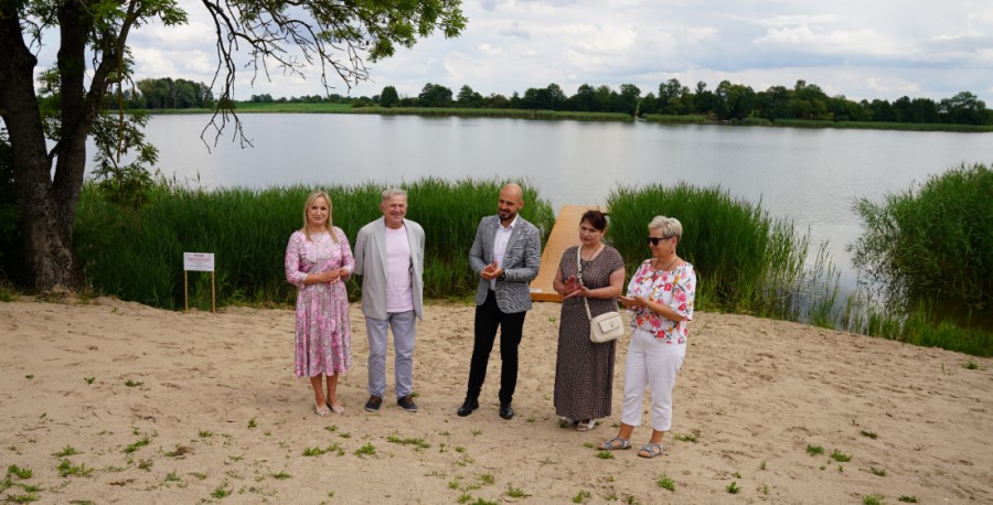 Pięć osób stoi na piaszczystym brzegu jeziora z pomostem, w tle trzciny i drzewa. Pogodny dzień, niebo częściowo zachmurzone. Grupa  w czasie  uroczystości plenerowej.
