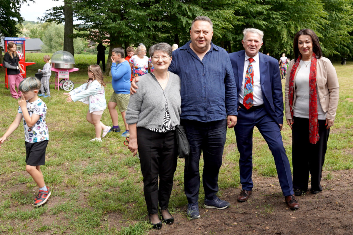 Grupa dorosłych i dzieci na pikniku na trawie. Na pierwszym planie cztery osoby pozują do zdjęcia, w tle wata cukrowa i inne atrakcje.