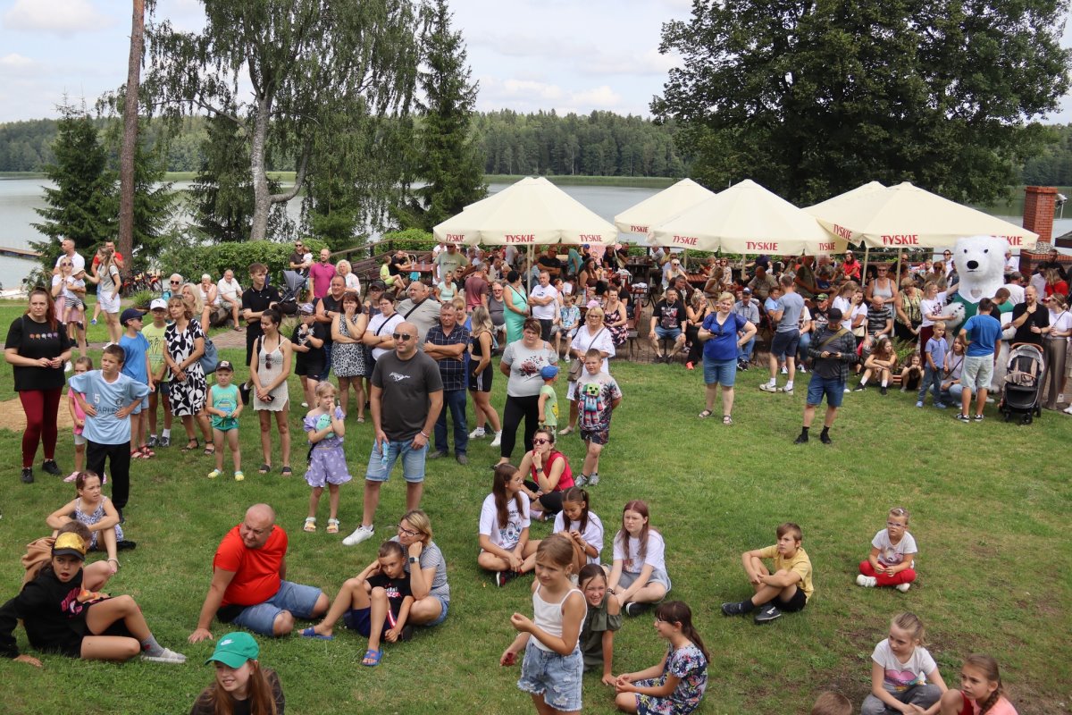 Duży tłum ludzi na festynie nad jeziorem. Dzieci siedzą na trawie, dorośli stoją i patrzą w jednym kierunku. W tle parasole „Tyskie”, las i jezioro. Obok maskotka – biały niedźwiedź. Letnia pogoda.