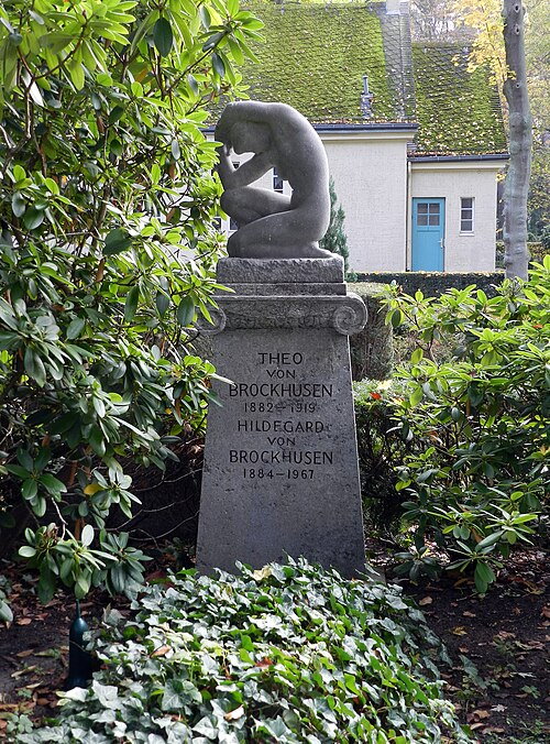 Nagrobek otoczony zielenią, z rzeźbą osoby w zamyślonej pozycji na szczycie. Na kamieniu widnieją nazwiska: Theo von Brockhusen (1882–1919) i Hildegard von Brockhusen (1884–1967).