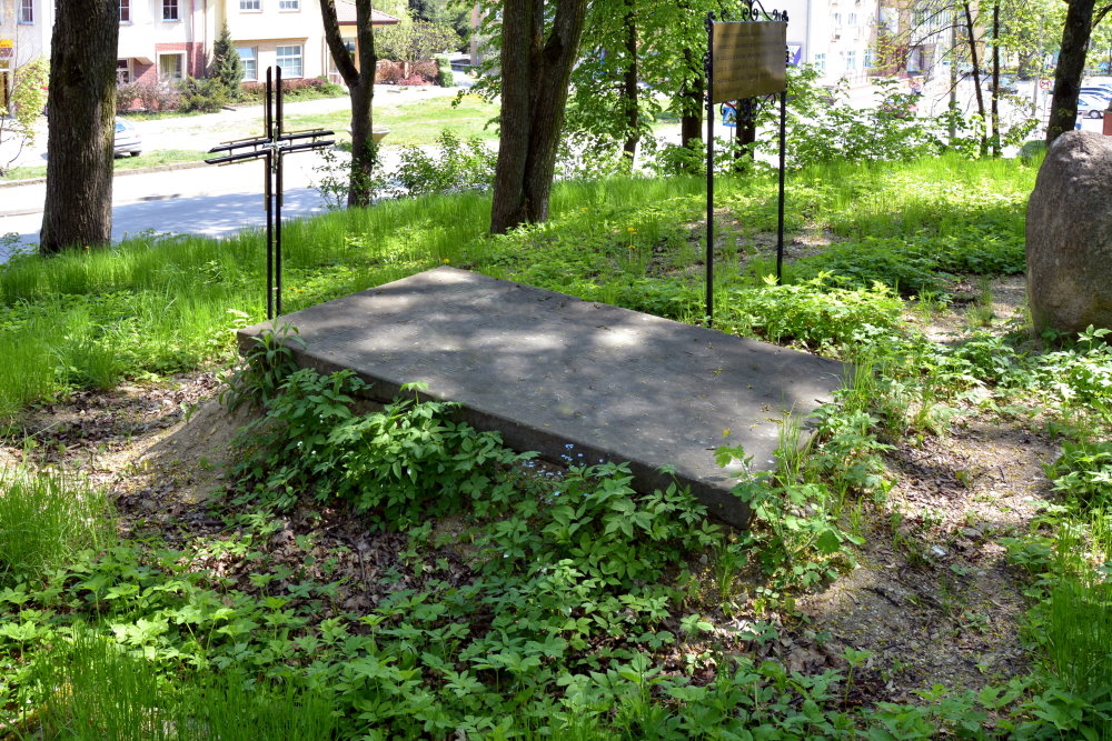Grób z betonową płytą wśród drzew i zieleni. Obok metalowy krzyż i tablica informacyjna. W tle ulica i budynki. Miejsce wygląda na ciche i zadbane.