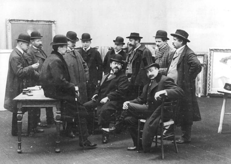 Czarno-białe zdjęcie grupy mężczyzn w melonikach i płaszczach, zgromadzonych w pomieszczeniu z obrazami opartymi o ścianę; część siedzi, część stoi, rozmawiają ze sobą.