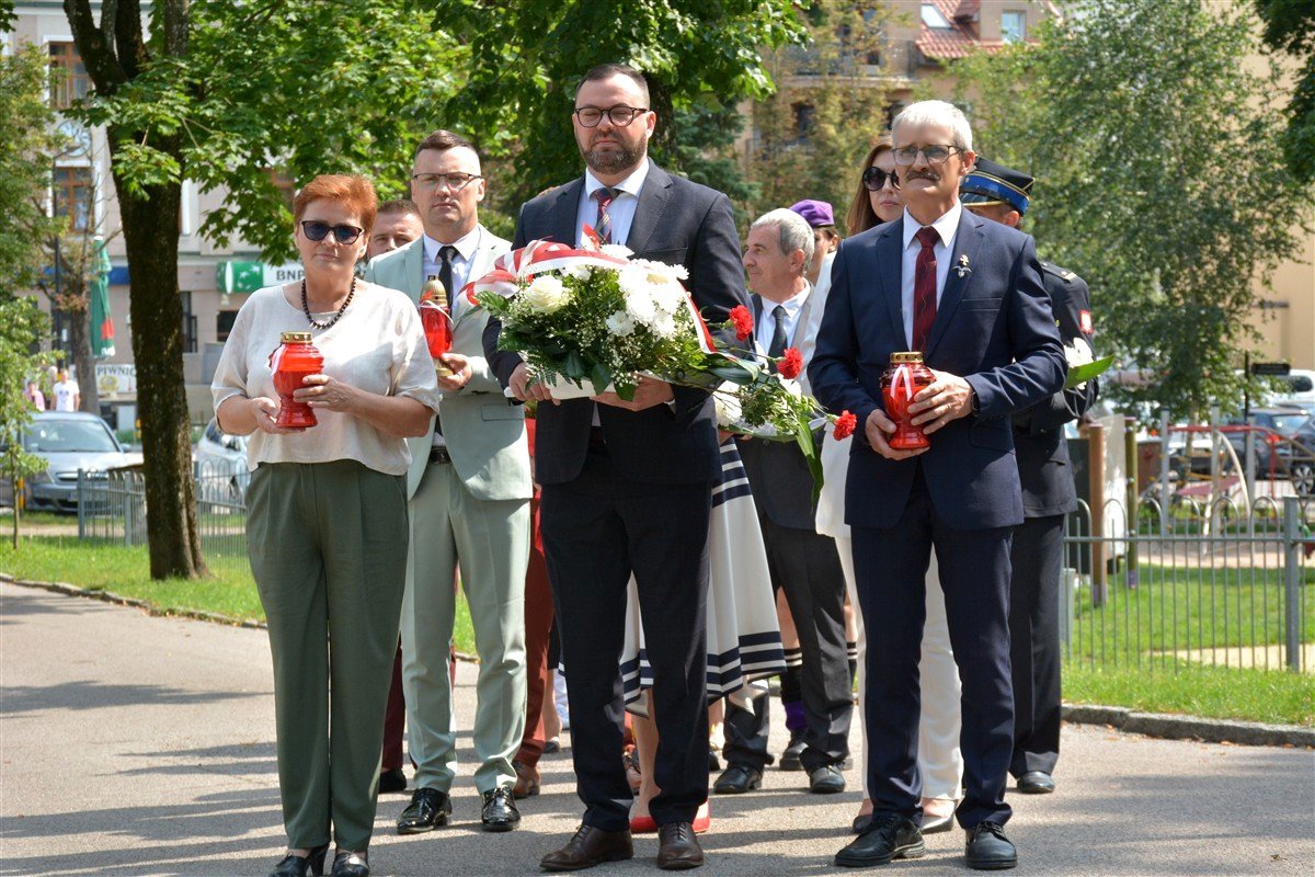 Grupa osób idzie w uroczystym pochodzie. Mężczyzna w środku trzyma wieniec z białych i czerwonych kwiatów, inni niosą czerwone znicze. W tle widać drzewa, budynki i zaparkowane samochody.