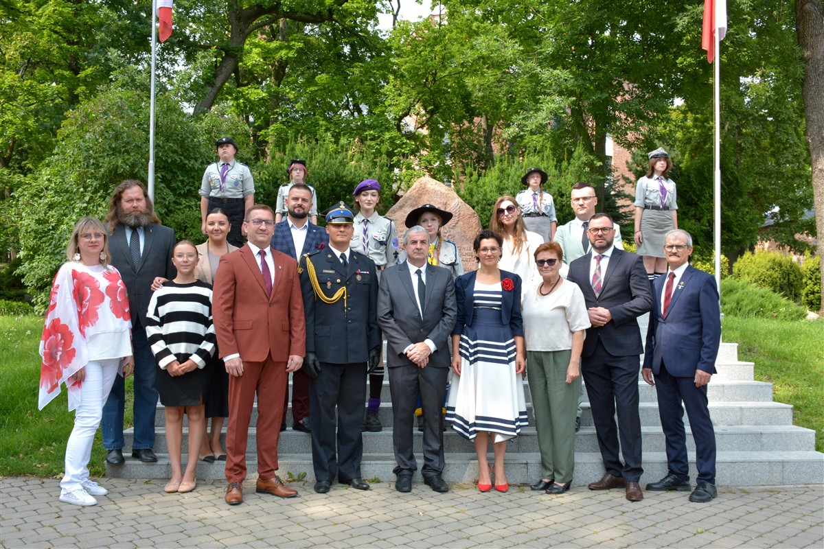Grupa osób, w tym harcerze, samorządowcy i przedstawiciele służb mundurowych, pozuje do zdjęcia przed pomnikiem. W tle drzewa i polskie flagi na masztach.