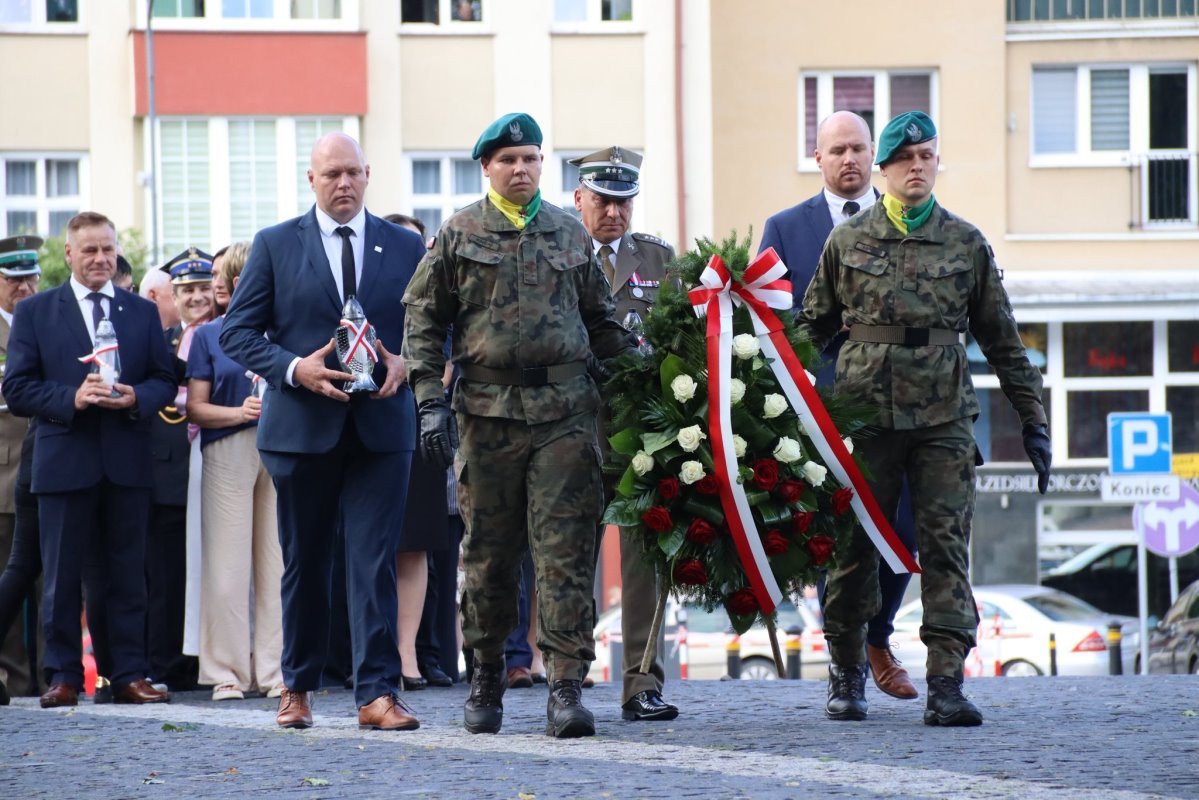 Uroczystość patriotyczna – dwaj żołnierze w mundurach niosą wieniec z biało-czerwonymi kwiatami, obok nich mężczyźni w garniturach trzymają znicze, w tle uczestnicy i budynki miejskie.