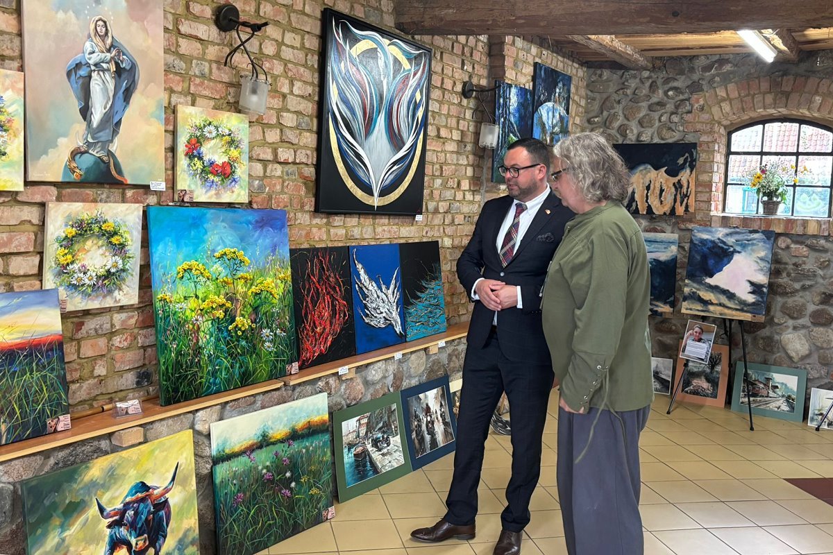 Dwóch mężczyzn ogląda obrazy w galerii sztuki z ceglanymi ścianami. Wokół nich wiszą i stoją kolorowe malowidła przedstawiające pejzaże, kwiaty, abstrakcje i postać religijną.