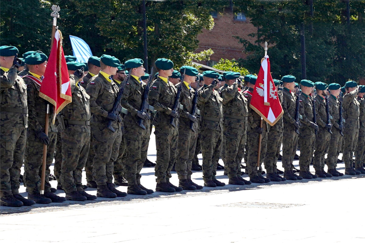 Żołnierze w mundurach polowych i zielonych beretach stoją w szeregu podczas uroczystości wojskowej. Część z nich salutuje, obok widoczne są sztandary z biało-czerwonymi barwami i orłem.