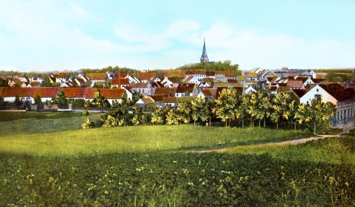 Widok miasta Olecko z początku XX wieku – kolorowa panorama z zielonymi polami, drzewami i zabudową, w tle widoczna wieża kościoła. 