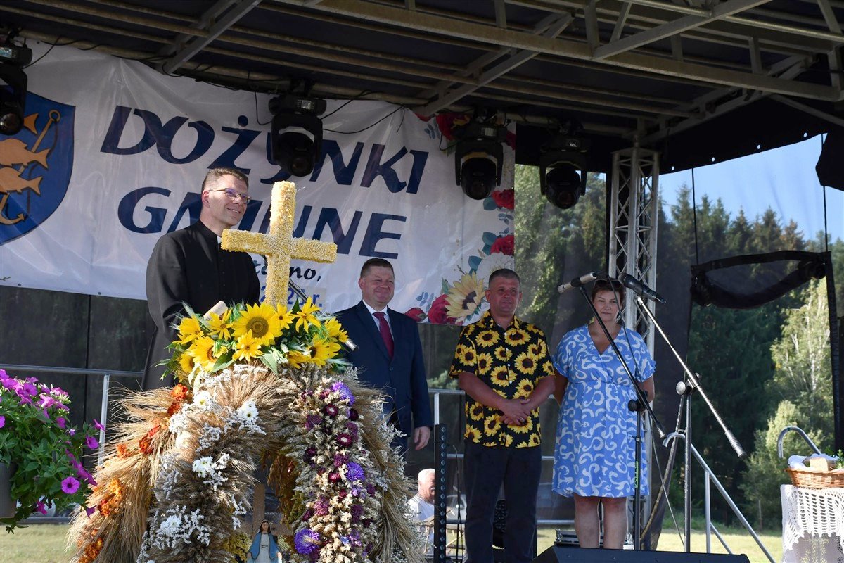 Ksiądz i przedstawiciele gminy stoją na scenie podczas dożynek. Przed nimi ustawiony jest wieniec dożynkowy z kłosów zbóż, kwiatów i żółtym krzyżem ozdobionym słonecznikami.