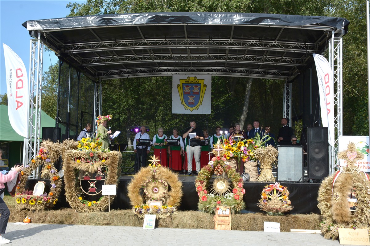 Scena podczas dożynek z zespołem śpiewaczym w strojach ludowych. Przed sceną ustawione są kolorowe wieńce dożynkowe z kwiatów, zbóż i symboli religijnych.
