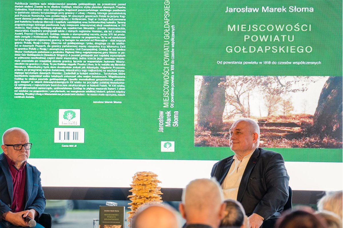 Dwaj mężczyźni siedzą na scenie podczas spotkania autorskiego. W tle widać wielki ekran z okładką książki „Miejscowości powiatu gołdapskiego” Jarosława Marka Słomy. Na stole stoi egzemplarz książki i tradycyjny sękacz.