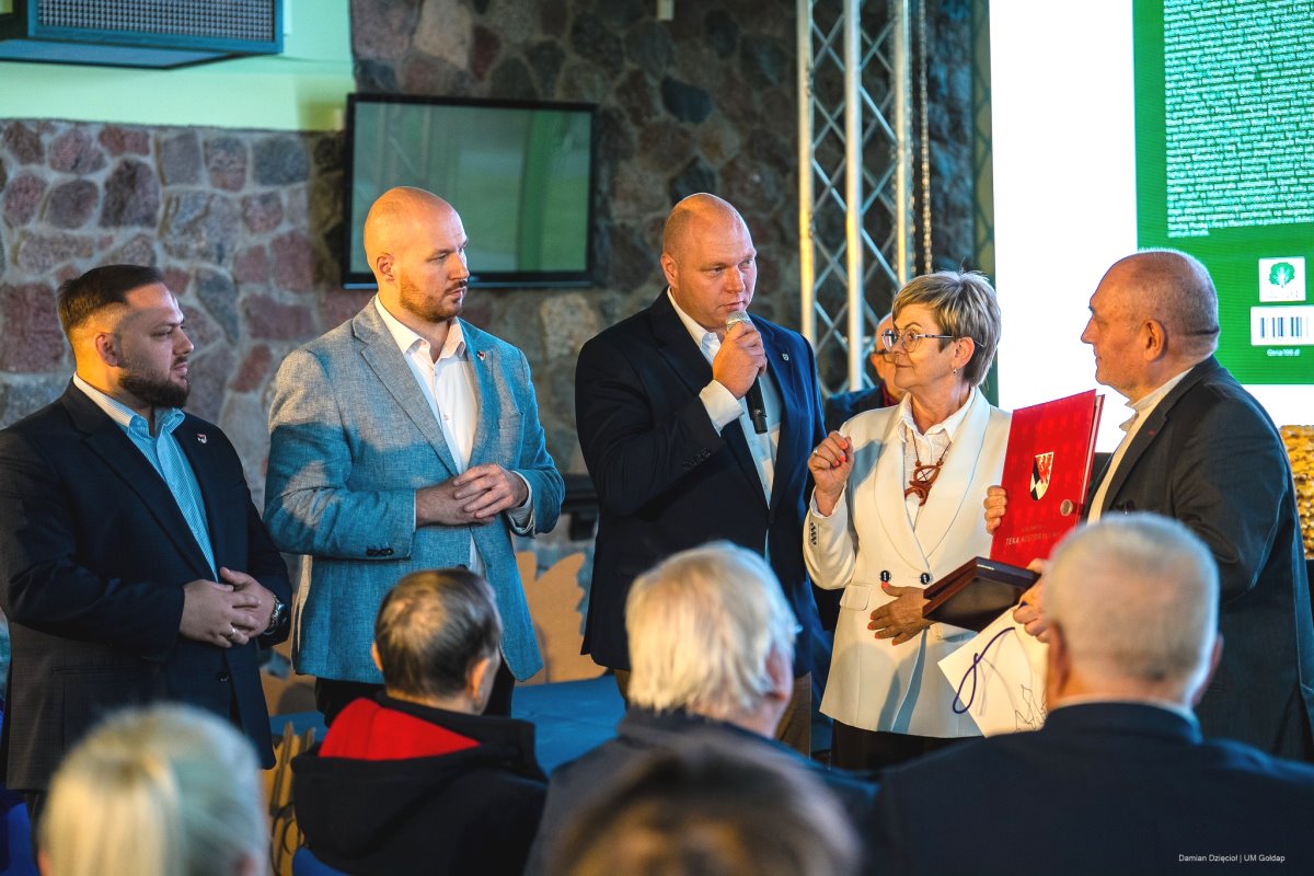 Grupa osób w eleganckich strojach stoi na scenie podczas oficjalnego wydarzenia. Jeden z mężczyzn mówi do mikrofonu, a kobieta wręcza starszemu mężczyźnie czerwoną teczkę z herbem. W tle widać publiczność i fragment ekranu.