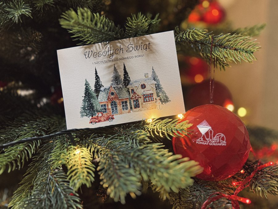 Gałązki choinki ozdobione czerwoną bombką i kartką świąteczną z zimowym pejzażem.