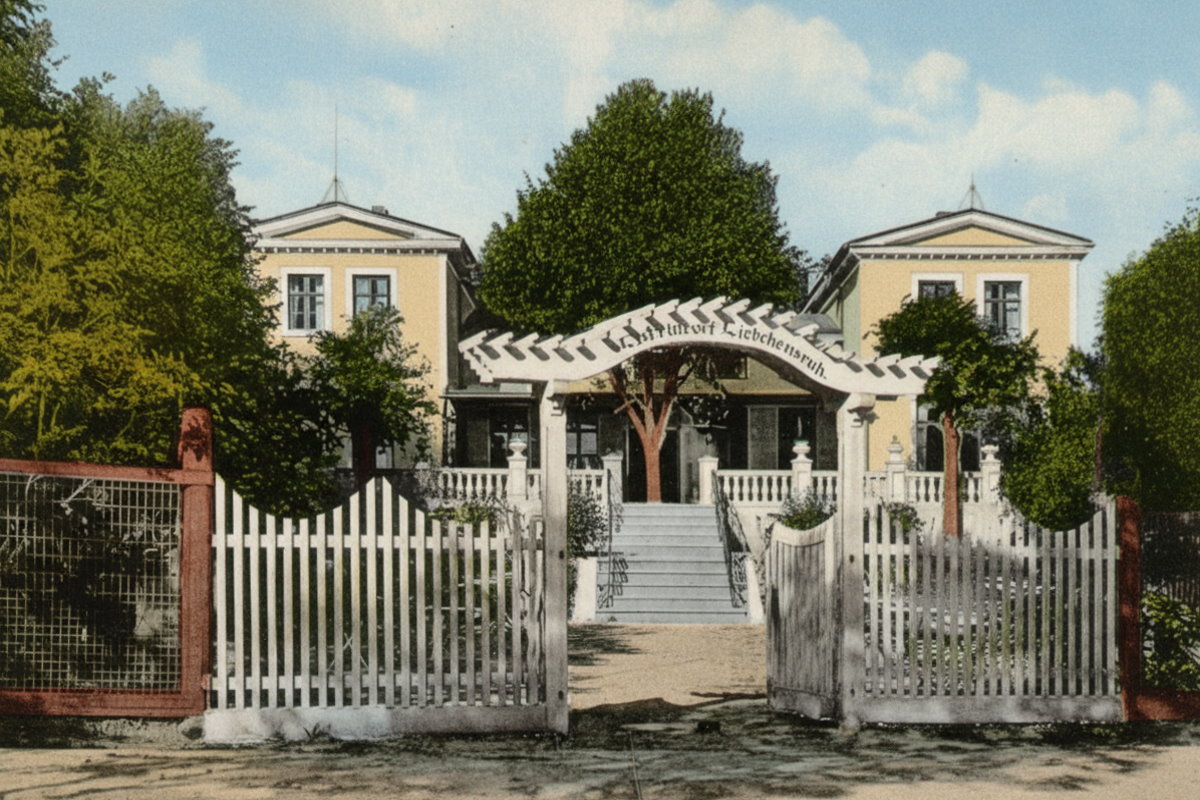 Elegancki budynek z kolumnowym wejściem i ozdobnym ogrodzeniem, otoczony zielenią, w spokojnej miejskiej okolicy.