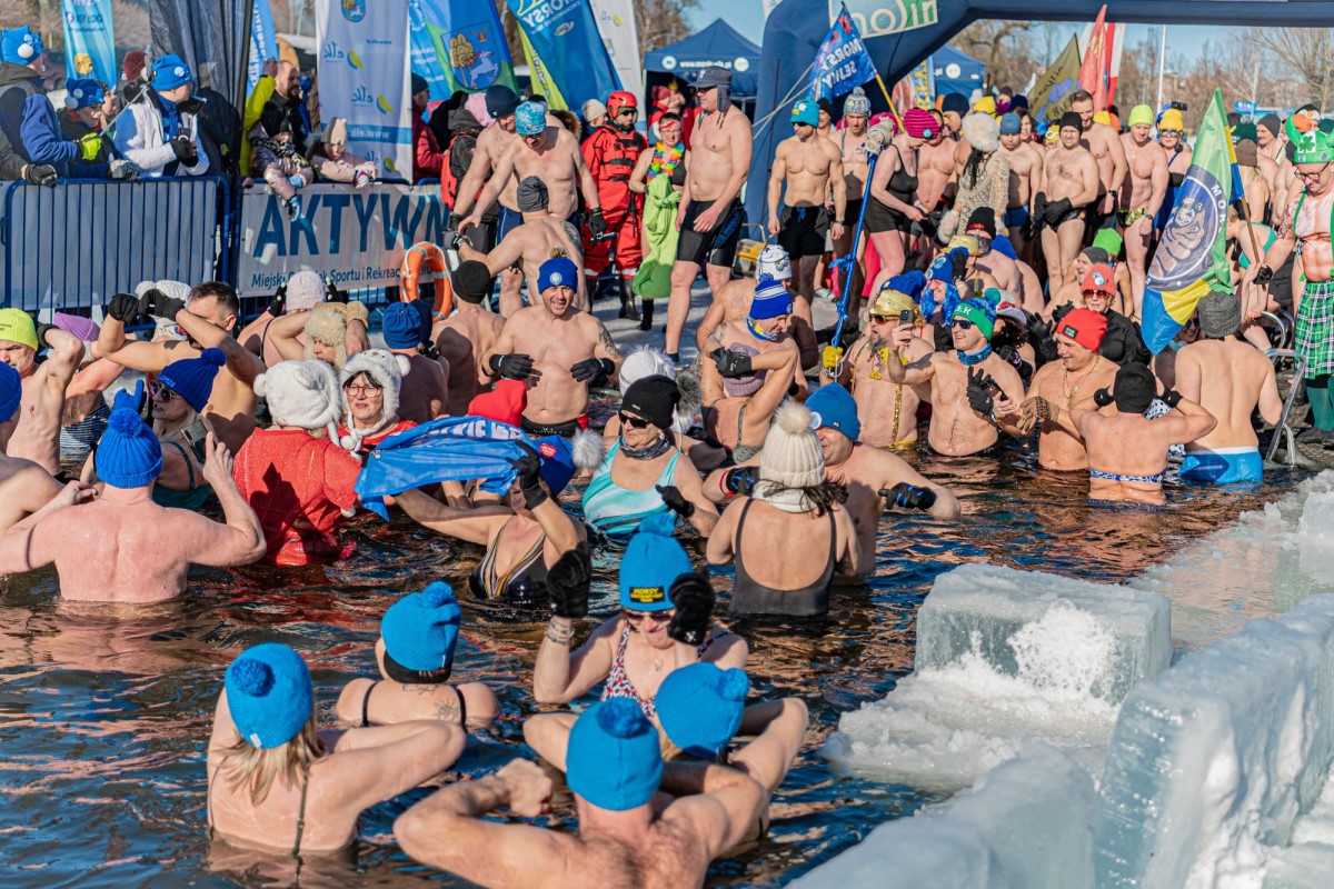 Tłum mors&oacute;w w czapkach kąpie się w przeręblu na jeziorze podczas zimowej imprezy, wok&oacute;ł widoczne flagi, ratownicy i widzowie na brzegu.