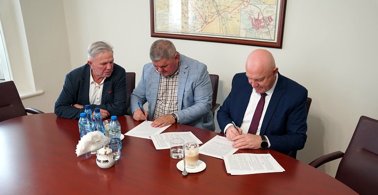 Trzech mężczyzn w garniturach siedzi przy stole konferencyjnym podczas podpisywania dokument&oacute;w; na stole stoją butelki wody, filiżanka kawy i rozłożone umowy.