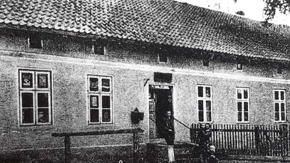 Archiwalne czarno-białe zdjęcie przedstawiające budynek mieszkalny z początku XX wieku. Przed wejściem stoi osoba, a przed domem znajduje się drewniane ogrodzenie i niewielki ogr&oacute;d.