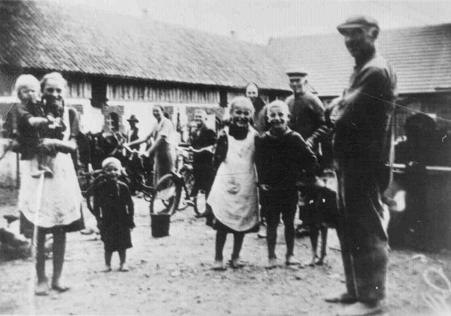 Historyczna czarno-biała fotografia przedstawiająca grupę mieszkańc&oacute;w zgromadzonych na podw&oacute;rzu wiejskim. W centrum stoi mężczyzna przemawiający lub rozmawiający z zebranymi.