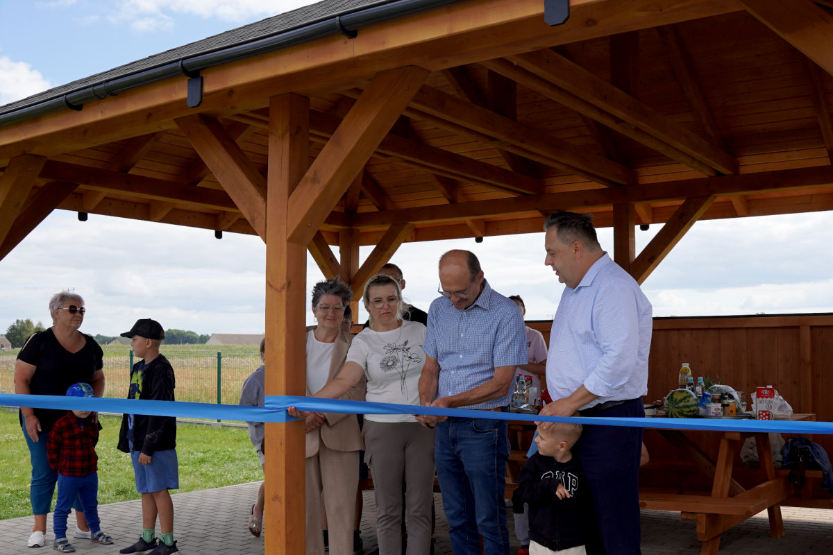 Grupa os&oacute;b podczas uroczystego przecięcia niebieskiej wstęgi pod drewnianą wiatą. W tle wiejska sceneria, st&oacute;ł z poczęstunkiem oraz dzieci i dorośli uczestnicy wydarzenia.
