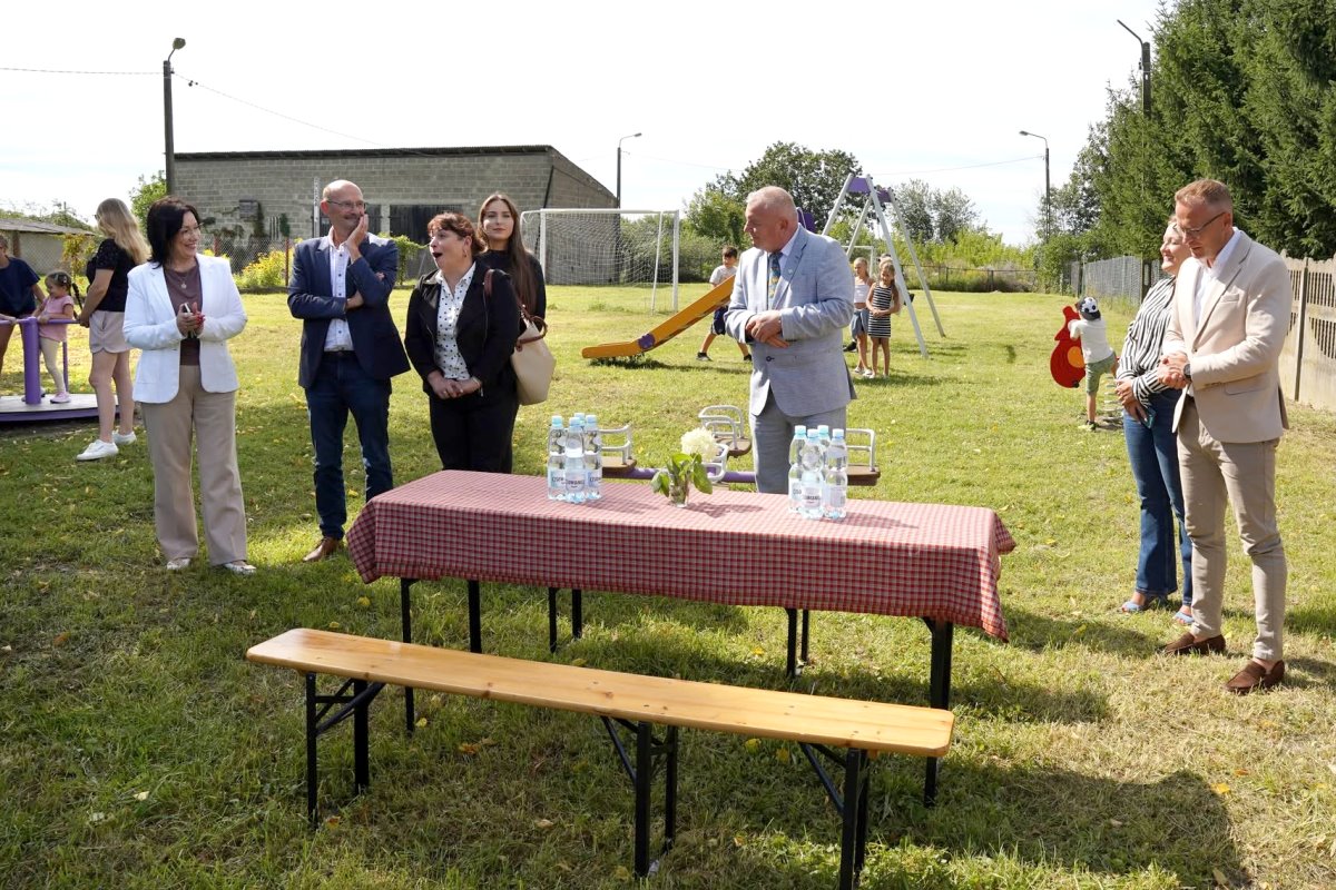 Uczestnicy uroczystego otwarcia placu zabaw stoją przy stole z poczęstunkiem na terenie rekreacyjnym.
